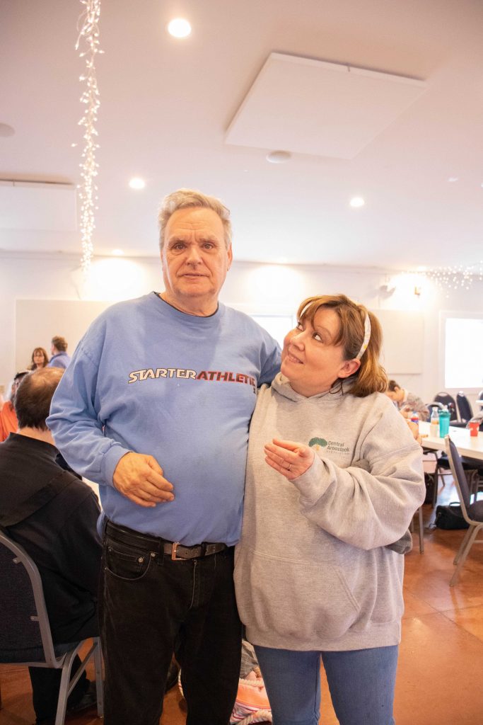 A woman with short red-brown hair wearing a grey sweatshirt looks up at a man with white hair who is wearing a blue long sleeved shirt